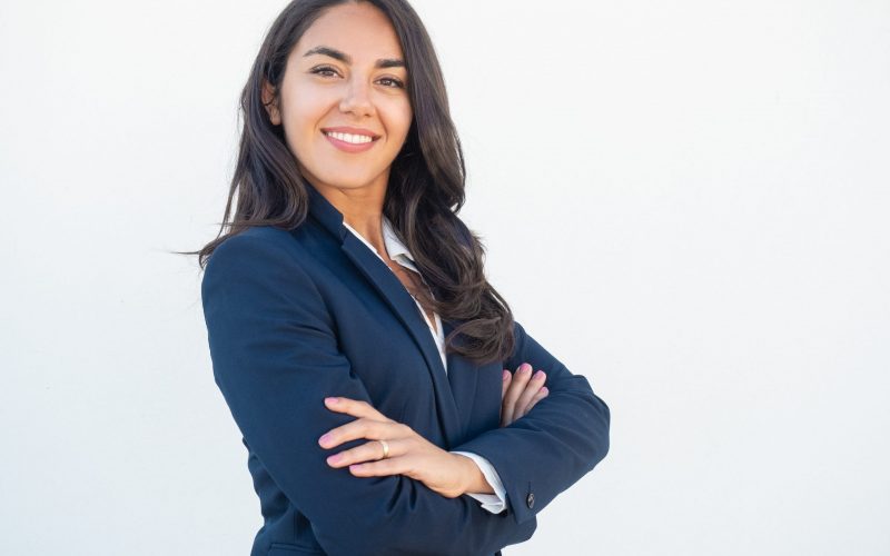 Smiling confident businesswoman posing with arms folded. Happy beautiful black haired young Latin woman in formal suit standing for camera over white studio background. Corporate portrait concept